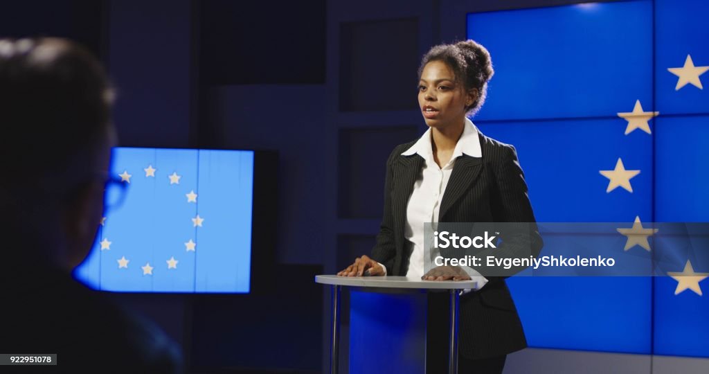 Female politician of EU having press conference Serious African-American representative of European Union answering questions of journalists on news conference in semilit studio with EU flag on screen. Women Stock Photo