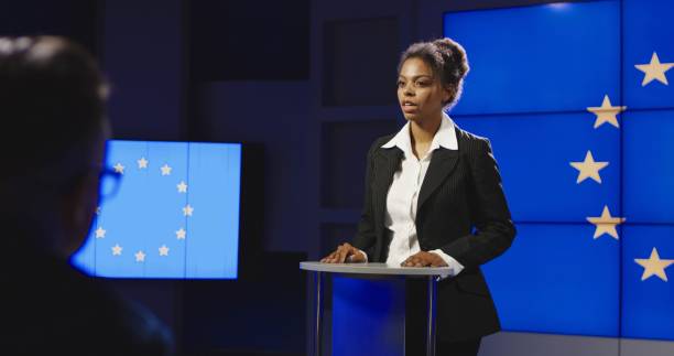 Female politician of EU having press conference Serious African-American representative of European Union answering questions of journalists on news conference in semilit studio with EU flag on screen. spokesperson stock pictures, royalty-free photos & images