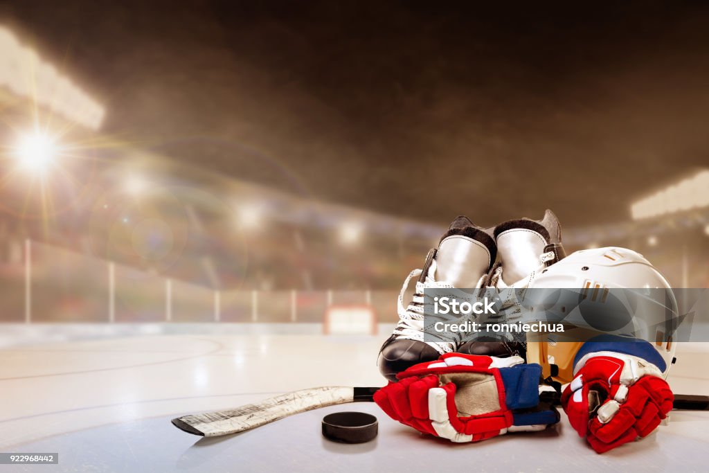 Outdoor Hockey Stadium With Equipment on Ice Ice hockey helmet, skates, stick and puck in brightly lit outdoor stadium with focus on foreground and shallow depth of field on background. Deliberate lens flare and copy space. Fictitious background arena created entirely in Photoshop. Hockey Stock Photo