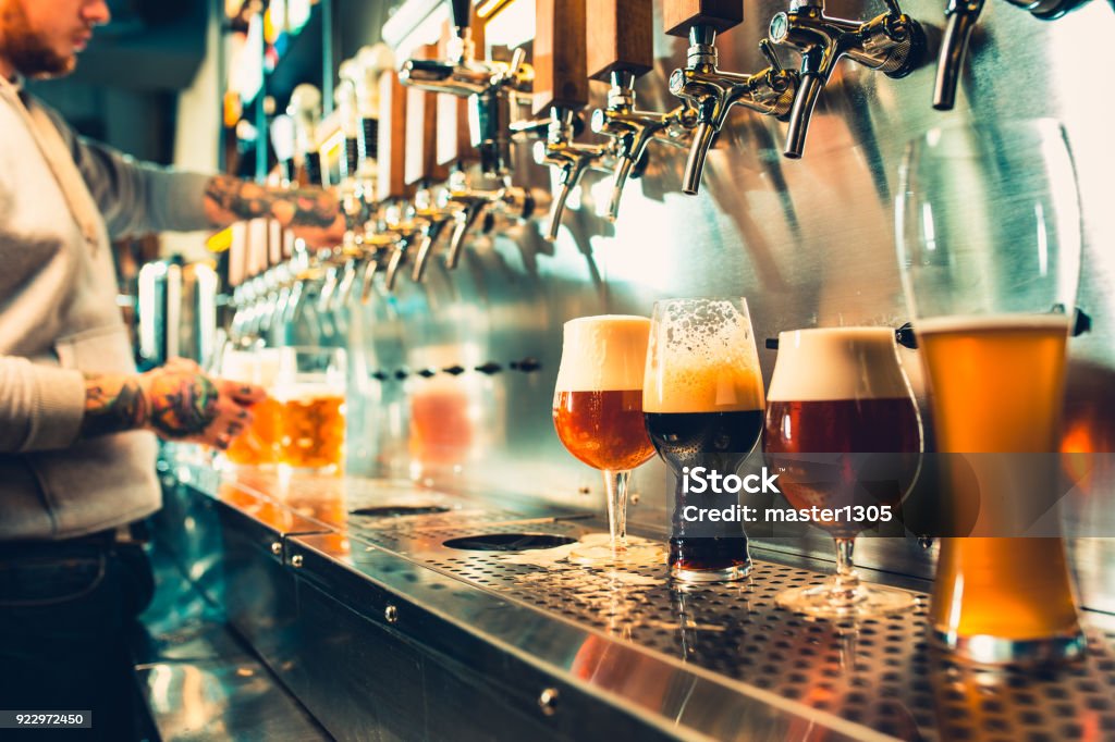 Glass of beer with barrel, bottle and fresh hops still-life We meet Beer Fest. Hand of bartender pouring a large lager beer in tap. Pouring beer for client. Side view of young bartender pouring beer while standing at the bar counter Beer - Alcohol Stock Photo