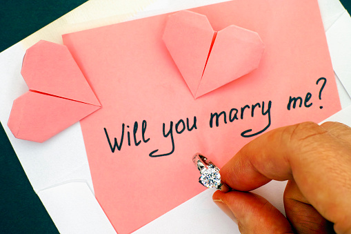 Person hand holding engagement ring. Letter with text Will you marry me? in envelope and two origami hearts on the table.