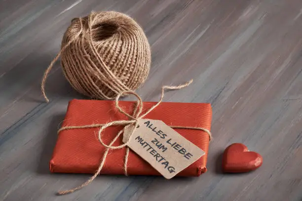 Wrapped gift tied up with cord, cardboard tag with text "Alles Liebe zum Muttertag" and a wooden heart on rustic wooden table. The text means "All the best for the Mother's Day" in German.