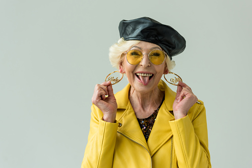 stylish senior woman in yellow leather jacket, earrings and yellow sunglasses, isolated on grey
