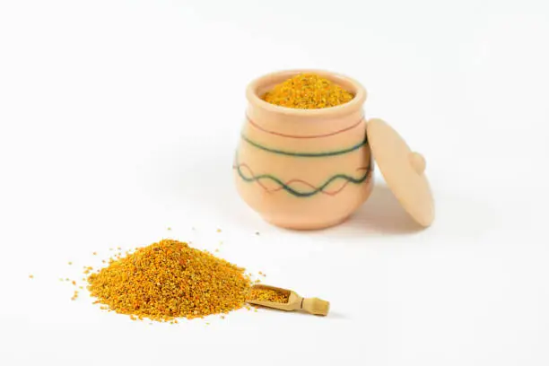 Pile of bee pollen and a little wooden scoop. In a blurry background, it is visible glazed clay pot full of pollen with a lid leaning against the pot.