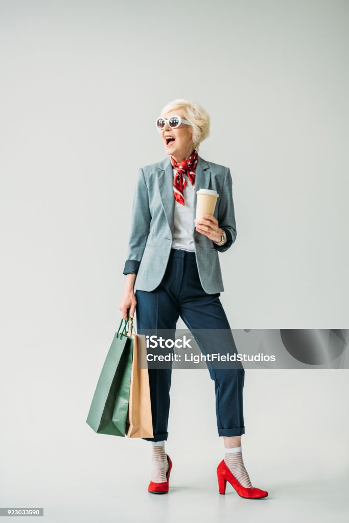 senior woman with shopping bags happy senior woman in jacket and sunglasses with shopping bags and coffee, isolated on grey Fashion Stock Photo