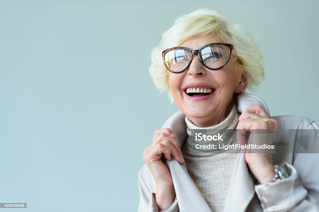 senior lady in eyeglasses beautiful senior lady in trench coat and eyeglasses, isolated on grey Senior Adult Stock Photo