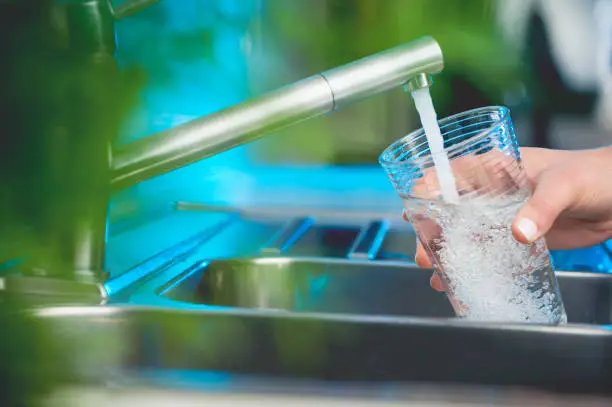 Woman filling a glass of water. She is using the faucet in the kitchen sink. There is a plant out of focus in the foreground. Close up with copy space.