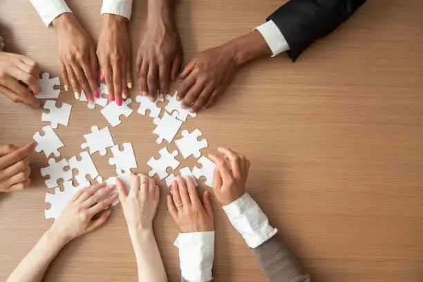 Hands of multi-ethnic team assembling jigsaw puzzle, multiracial group of black and white people joining pieces at desk, successful teamwork concept, help and support in business, close up top view