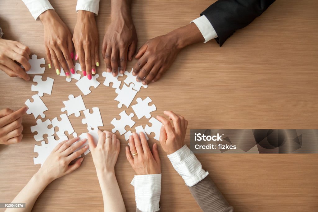 Hands of multi-ethnic business team assembling jigsaw puzzle, top view Hands of multi-ethnic team assembling jigsaw puzzle, multiracial group of black and white people joining pieces at desk, successful teamwork concept, help and support in business, close up top view Multiracial Group Stock Photo