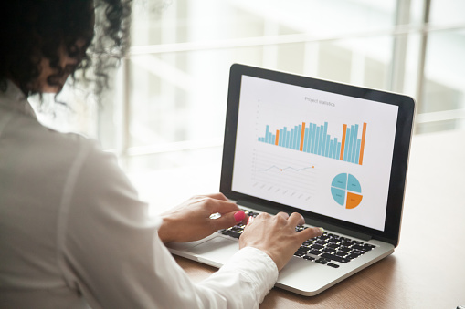 African businesswoman analyzing statistics on laptop screen, working with financial graphs charts online, using business software for data analysis and project management concept, rear close up view