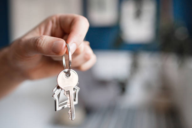 istock Mortgage concept. Men hand holding key with house shaped keychain. Modern light lobby interior. Real estate, moving home or renting property. 923061846