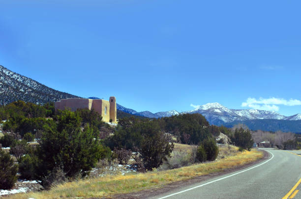 Road curves around the San Francisco de Asis Church near Golden, New Mexico.  Building sits on hill in front of mountains.