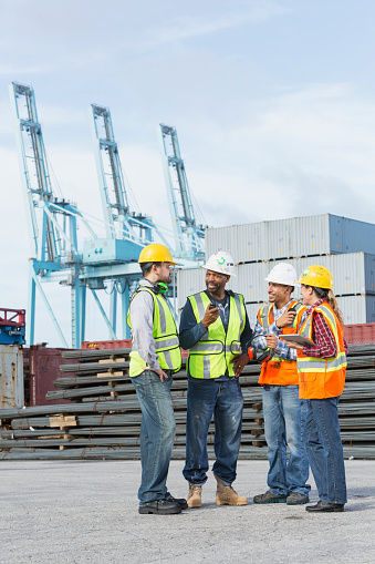 A group of three multi-ethnic men and a woman wearing safety vests and hardhats, working at a shipping port with cargo containers and cranes in the background.  The mature woman in her 40s is holding a digital tablet.