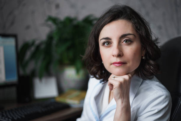 woman doctor with an attentive serious look in white medical robe sitting at a table. on the table books, a computer monitor and a green plant. the horizontal frame. the background is not in focus. - portrait doctor paramedic professional occupation imagens e fotografias de stock