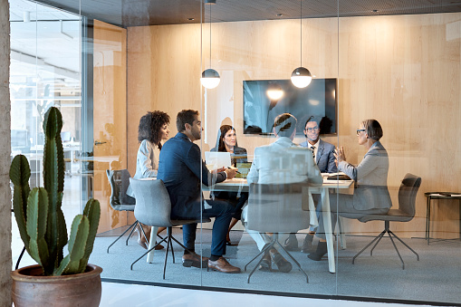 Senior businesswoman explaining strategy with colleagues in board room. Multi-ethnic professionals are discussing while sitting at desk in office. They are seen through glass.