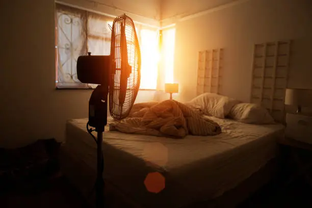 Electric fan in-front of an unmade bed with light coming through a window