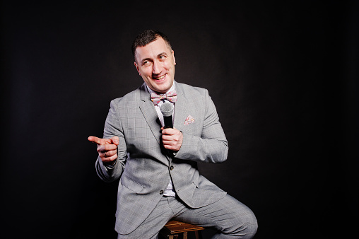 Handsome man in gray suit with microphone against black background on studio sitting on chair.  Laughs face of toastmaster and showman.