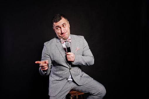 Handsome man in gray suit with microphone against black background on studio sitting on chair. Toastmaster and showman.