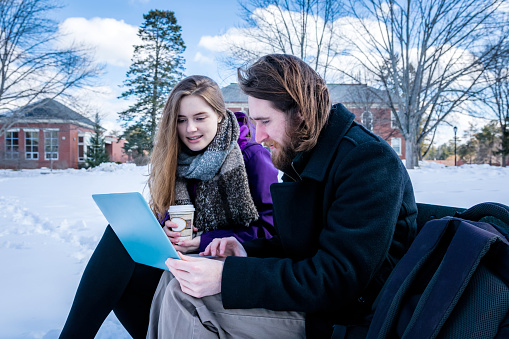 University students operating a laptop on a bench in the center of a snow filled field.