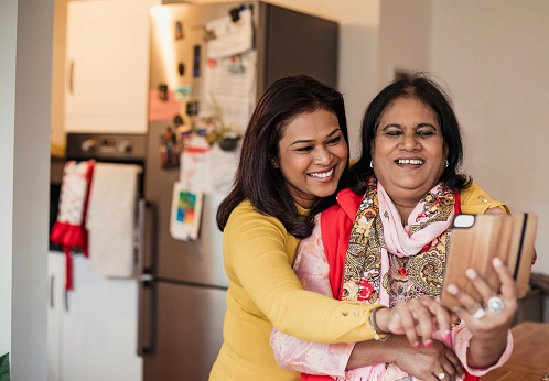 Two women take a selfie in the kitchen. The senior woman is wearing a duppatta.