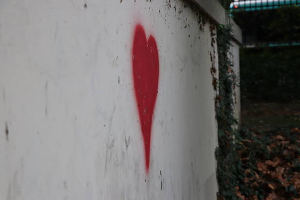 A vibrant red heart is painted or illuminated behind a chain-link fence, partially obscured by lush green foliage in the foreground. The warm glow of the heart contrasts with the cool, metallic lines of the fence, creating a visually interesting interplay of textures and emotions. This image evokes feelings of love, hope, and resilience, even when faced with barriers. It could represent hidden beauty, unexpected connections, or a message of affection peeking through.