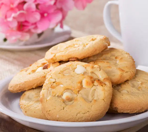 Closeup of a pile of macadamia nut cookies on a plate with coffee cup and flowers in background