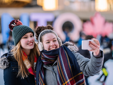 Young Caucasian teenage women taking selfies on the skating rink in downtown area of big city in North America. Both smiling, dressed in heavy winter clothes. In the background, unrecognizable people skating, neon signs of the city.