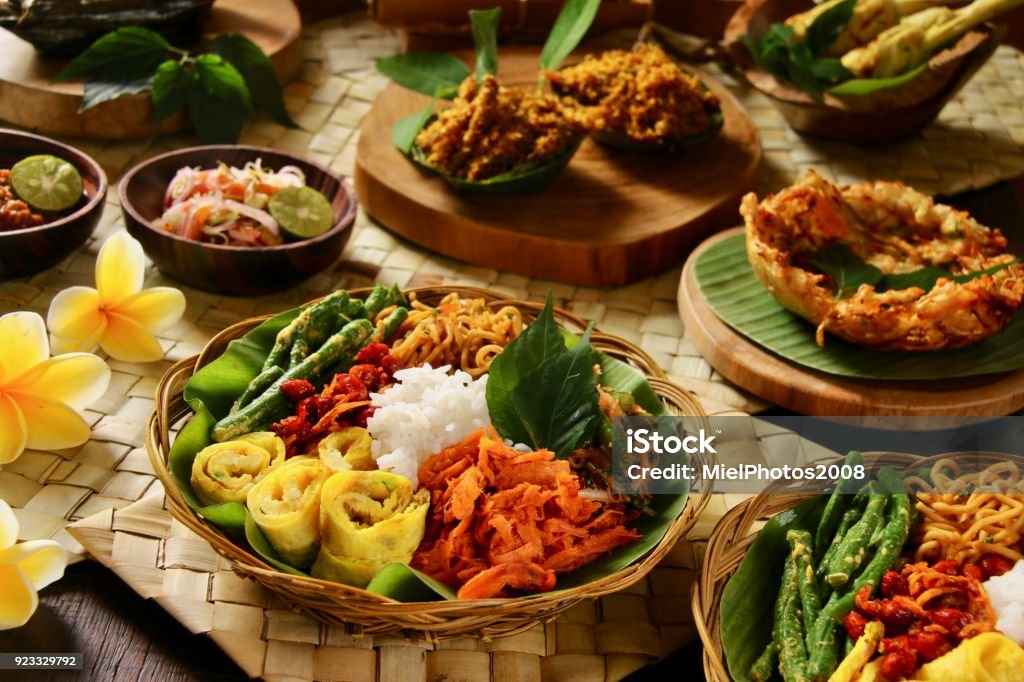 Nasi Campur Bali, a Popular Balinese Meal of Rice with Variety of Side Dishes Nasi Campur Bali, a popular traditional Balinese rice dish of steamed rice served with variety side dishes. Some of the side dishes are served together with the rice on the plate; while there are several more as optional extras. The complete rice meal is plated on a woven plate (basket) lined with banana leaf. These pre-plated meals are arranged on the table among the optional extra side dishes. Several frangipani flowers are used as table decoration. Food Stock Photo