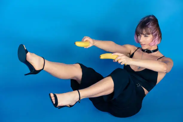 Photo of stylish girl sitting and holding bananas in hands in front of her isolated on blue