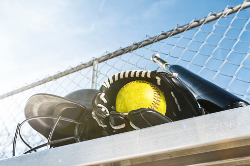 Looking up at a worn yellow softball in a black leather glove with a black batting helmet and a black aluminum bat on an aluminum bench with a chainlink fence and blue sky in the background with sun flare.