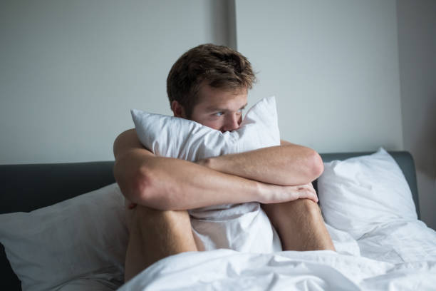 Ill caucasian man sitting on his bed. He is holding pilow because he is nervous. Concept of mental health problem