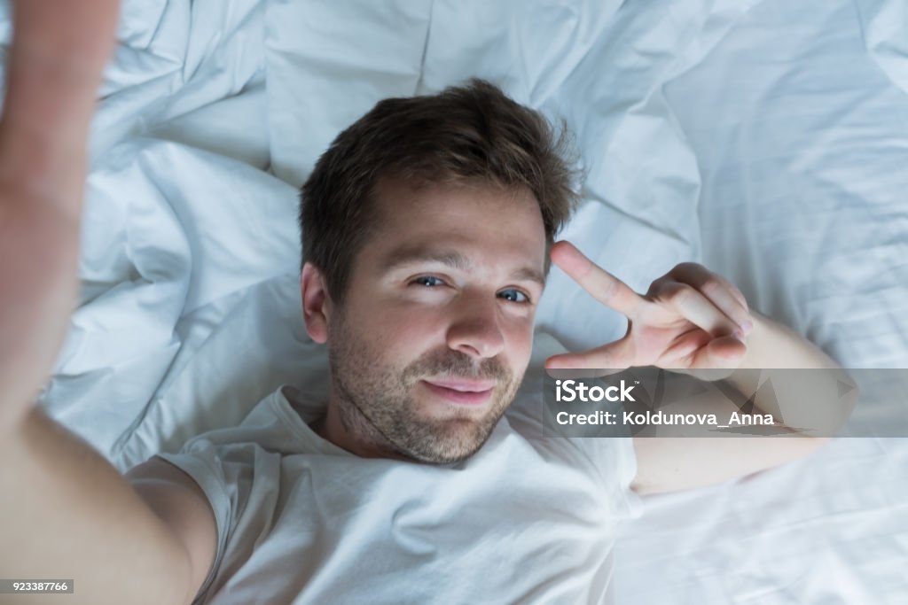 handsome man in white shirt taking a selfie in bed handsome man in white shirt taking a selfie in bed saying hello to his girlfriend in the morning Selfie Stock Photo