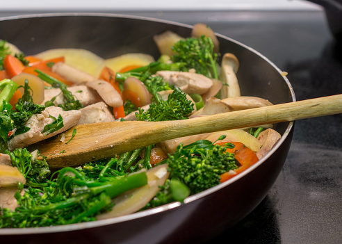 Close-up and side view of wooden spoon stirring chicken and vegetables in cast iron skillet - Stir frying