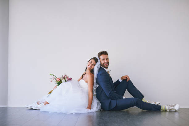 Studio shot of a newly married young couple sitting back to back on the floor against a gray background