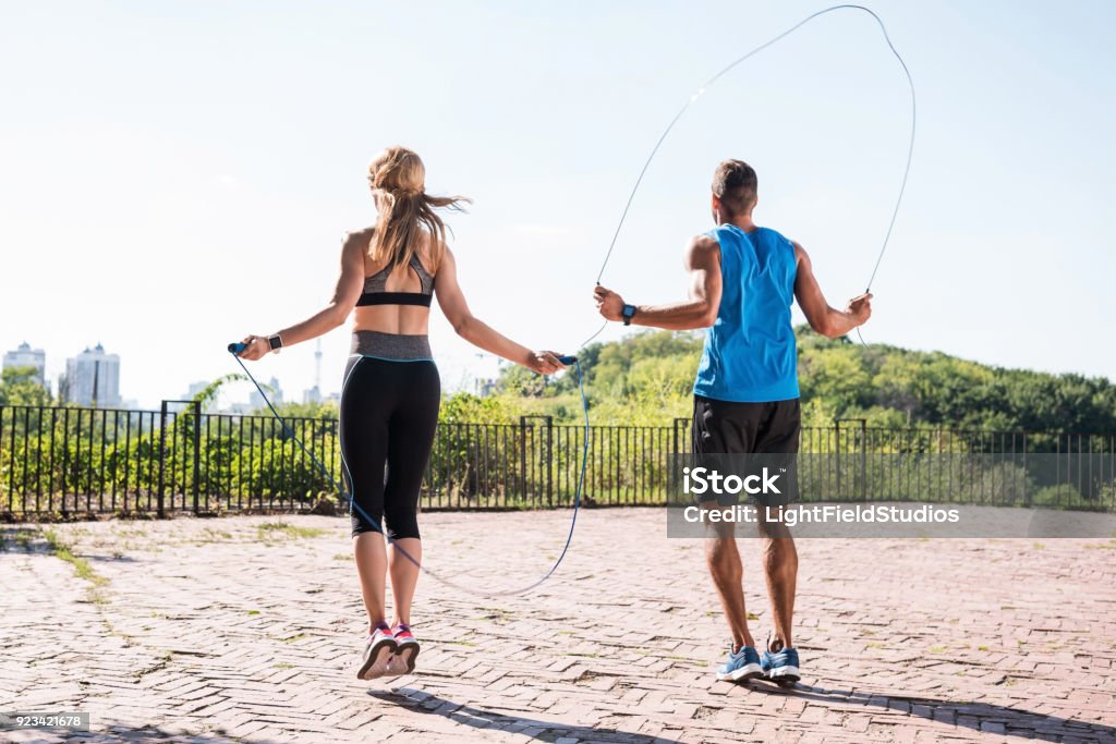 jumping on skipping ropes back view of athletic sportswoman and sportsman jumping on skipping ropes in park Jumping Rope Stock Photo