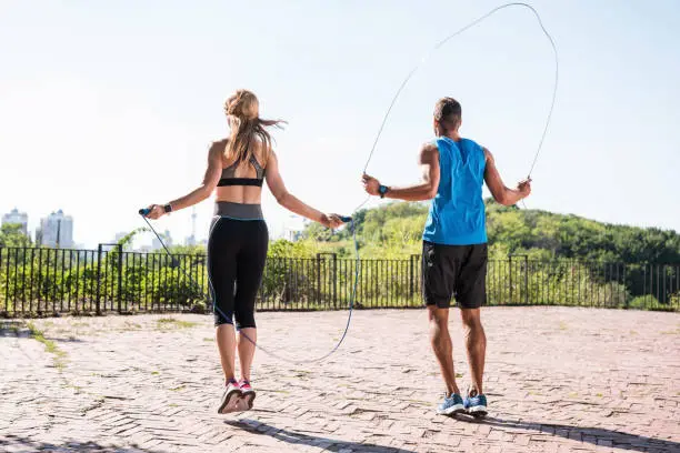 back view of athletic sportswoman and sportsman jumping on skipping ropes in park