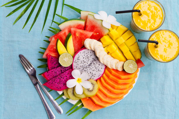 istock Tropical fruits assortment on a white plate with palm tree leaf with mango smoothie, textile blue background. Top view. Copy space 923447824