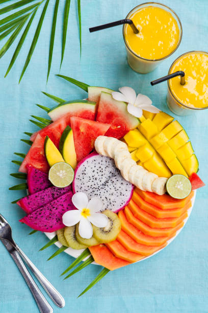 istock Tropical fruits assortment on a white plate with palm tree leaf with mango smoothie, textile blue background. Top view. Copy space 923448032