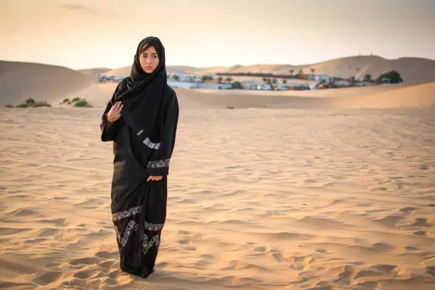 Photo of Full length portrait of Arabic woman in traditional black clothes standing in the desert in front of Bedouin village.