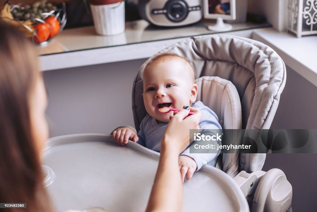 Mom feeds the baby from the spoon in the kitchen. A child is sitting in a child's chair. Baby - Human Age Stock Photo