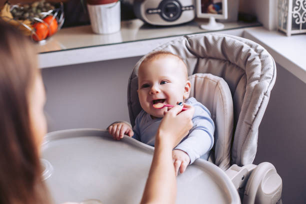 mom feeds the baby from the spoon in the kitchen. a child is sitting in a child's chair. - mother enjoyment built structure human head imagens e fotografias de stock
