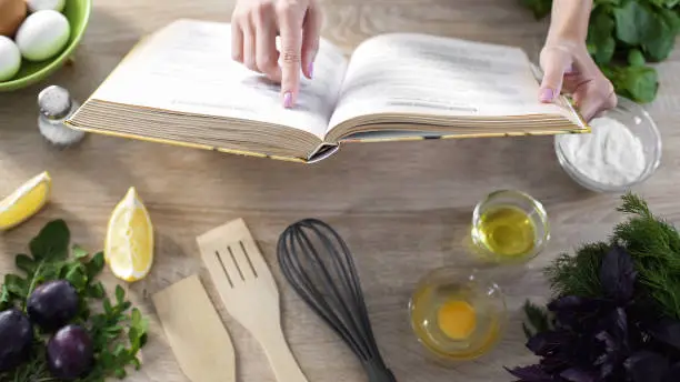Photo of Lady reading pizza recipe in culinary book at home with kitchenware on table
