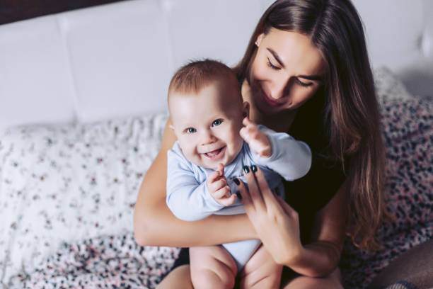 junge mutter mit dem kleinen kind sitzen auf einem bett und lächeln - offspring child toothy smile beautiful stock-fotos und bilder