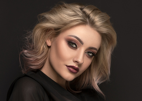 Delicate blonde blonde lady with glamour makeup posing in elegant style, looking at camera. Studio shot, dark background. Caucasian woman.