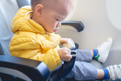 Cute little Asian 20 months / 1 year old toddler baby boy child wearing & fasten seat belts while sitting on airplane seat. Safety measures on board. Precautions on plane during flight for kid