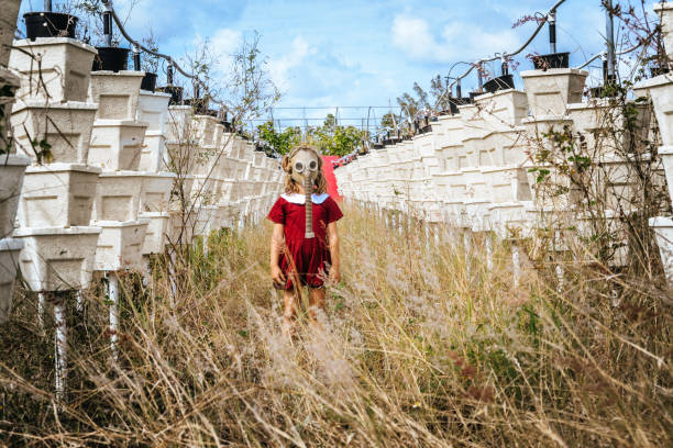 Young child in a vintage looking photograph stands in an empty field of overgrown weeds and empty vertical gardens wearing a gas mask. Commentary on pollution, pesticides, and a dystopian future