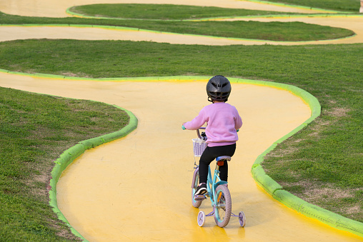 Back view of 4 years old girl riding on a bicycle with training wheels at the public park. Toddler cycling on yellow track.