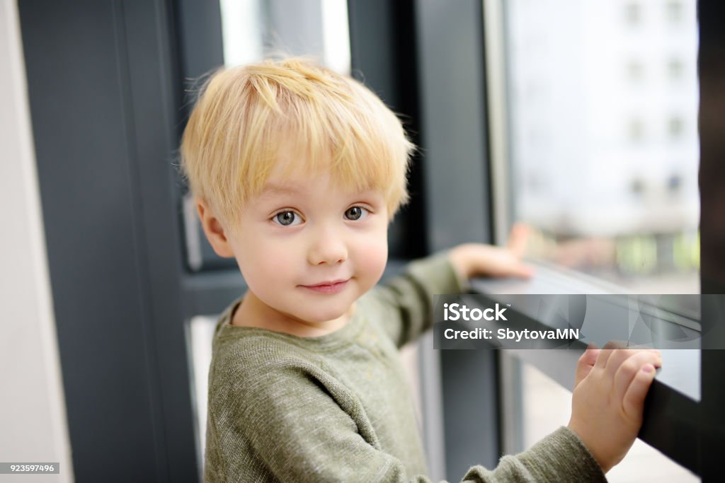 Lovely little boy sitting on the window near panoramic window and looking outside Lovely little boy sitting on the window near panoramic window and looking outside. Portrait of cute child with blonde hair Blond Hair Stock Photo