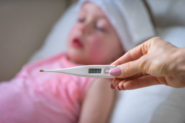 Sick child with high fever laying in bed and  holding thermometer.  Compress on forehead. stock photo