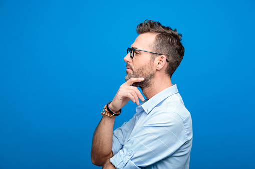 Summer portrait of confident, handsome man wearing blue shirt and glasses, looking away with hand on chin. Side view. Studio shot, blue background.